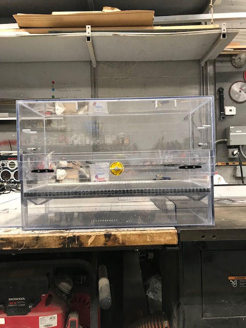 A clear, rectangular acrylic display case sits on a workbench in a cluttered workshop.
