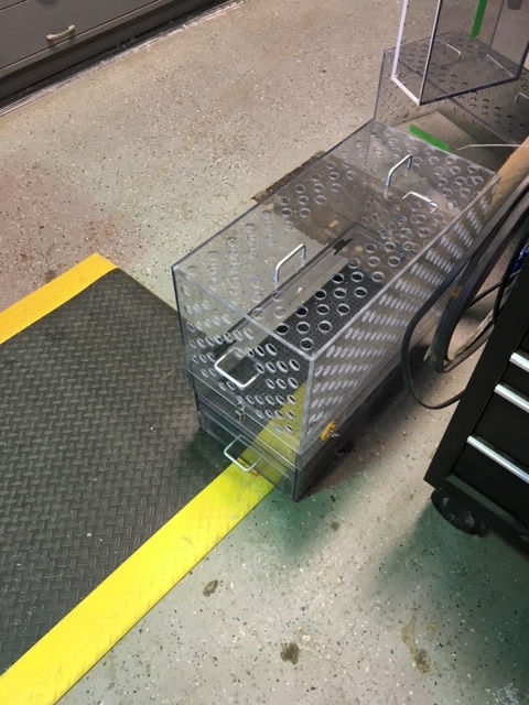 Clear, rectangular storage organizers with a grid-patterned lid and metal handles, sitting on a workshop floor.