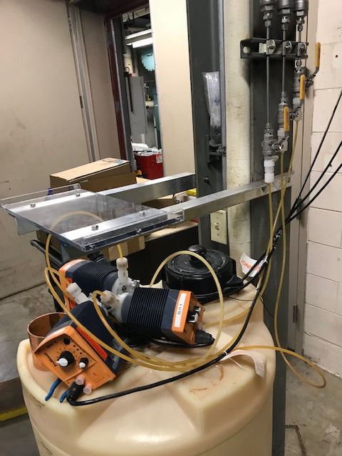 Chemical metering pumps mounted on a plastic storage tank, with tubing connected to a metal rack in an industrial room.