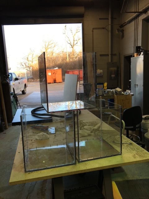 Two clear acrylic boxes, one stacked on top of the other, sitting on a wooden workbench in a workshop.