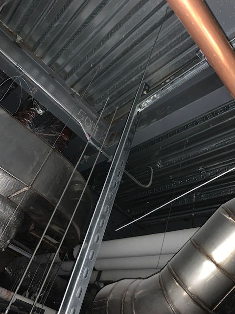 A view looking up at a commercial ceiling with exposed metal decking, support beams, pipes, and metal struts.