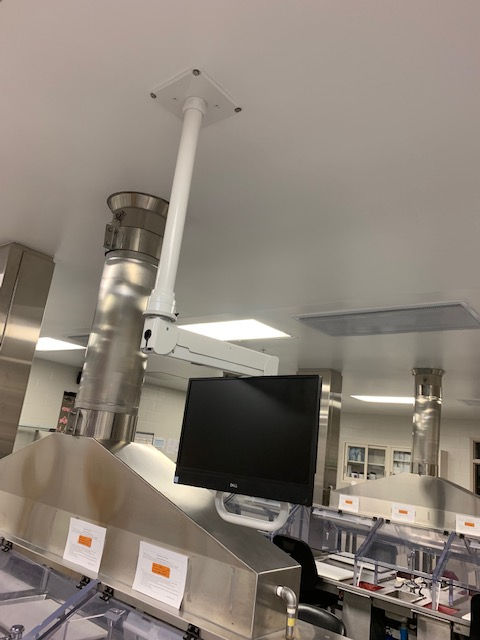 A ceiling-mounted computer monitor arm and screen positioned above stainless steel laboratory equipment in a bright room.