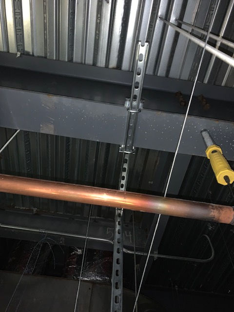 A copper pipe runs horizontally in front of a vertical metal strut hanging from a ribbed metal ceiling above a steel beam.