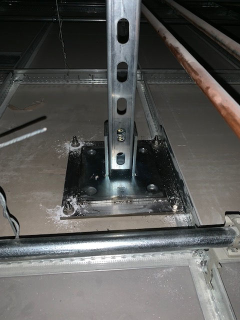 A metal vertical support post mounted to a floor using a square steel base plate with four bolted corners.