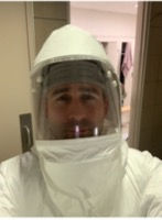 A person wearing a white hooded protective suit with a clear face shield, standing in an indoor setting.