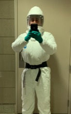 Person in a white protective suit with a clear face shield and green gloves taking a mirror selfie indoors.