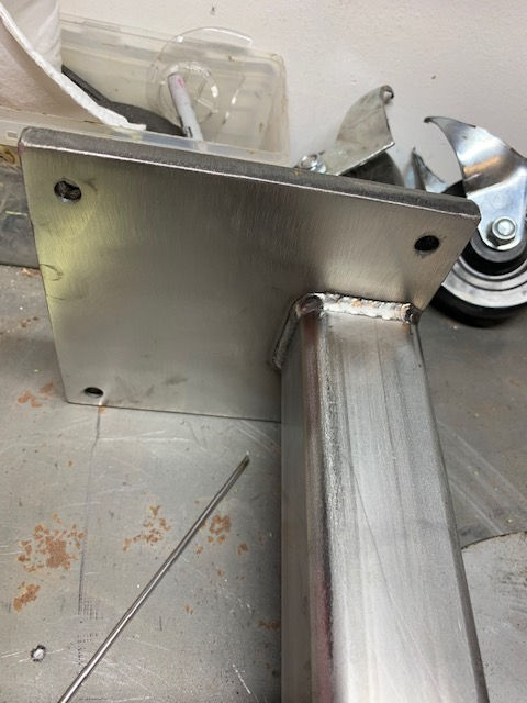 A square metal plate welded to a vertical rectangular steel post sitting on a workbench with casters in the background.