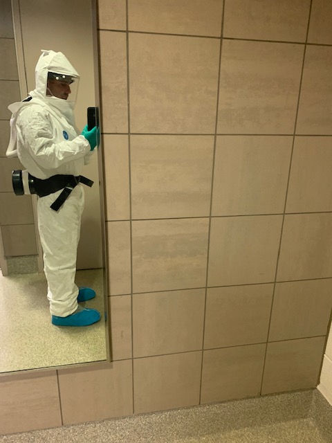A person wearing a full white hazardous materials suit with a respirator mask, taking a selfie in a tiled room mirror.