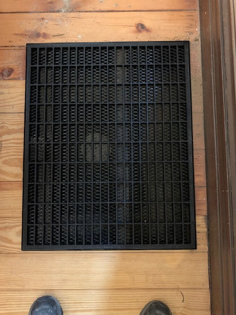 A black, rectangular rubber floor mat with a grid pattern, lying on a light wood floor.