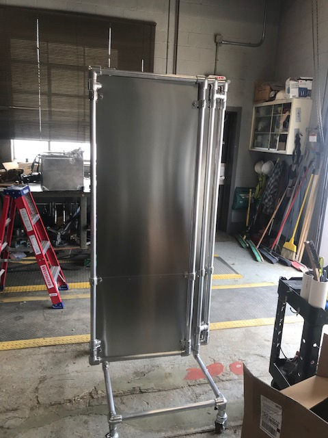 A tall, stainless-steel partition with a metal frame and caster wheels stands in a cluttered workshop.