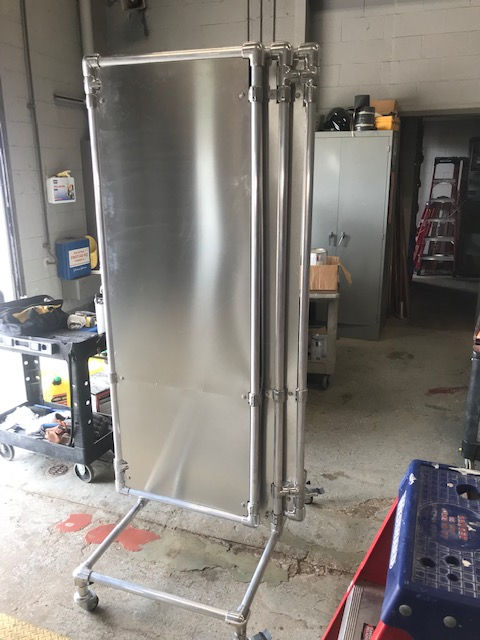 A tall, metallic portable folding partition on wheels stands in an industrial garage setting.