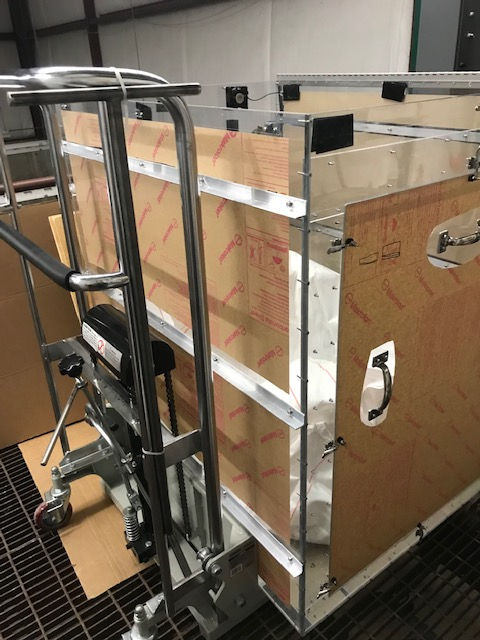 A transport cart loaded with a large box protected by clear acrylic panels and metal framing on a warehouse floor.