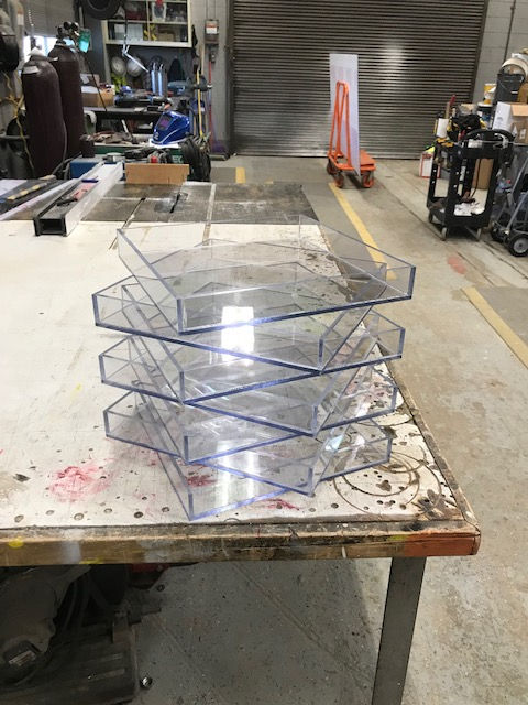 A stack of five clear, square acrylic trays sitting on a workbench in a cluttered workshop.