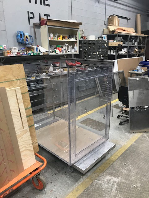 A clear, rectangular acrylic enclosure sits on a metal base in a workshop, near a stack of plywood on a cart.