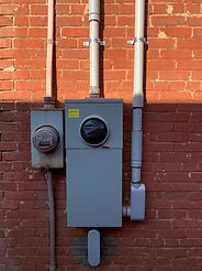 Gray electrical service equipment, including two meters and conduit piping, mounted on a red brick exterior wall.