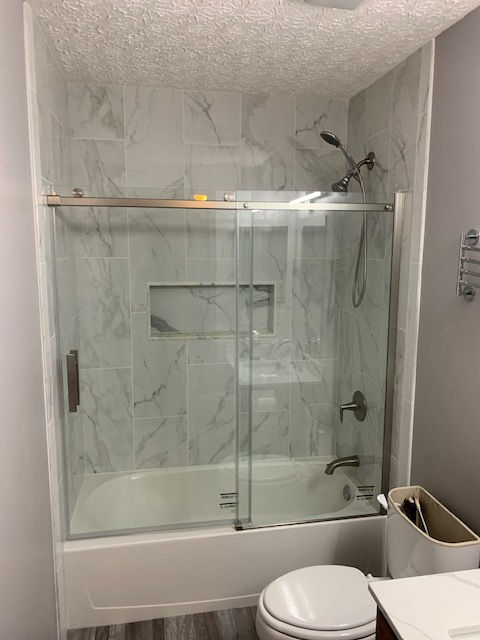 A bathroom featuring a bathtub with a glass sliding door, marble-patterned tile walls, a built-in niche, and a showerhead.