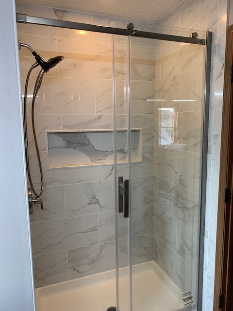 A glass shower enclosure with white marble-patterned tile, a built-in recessed shelf, and a handheld shower head.