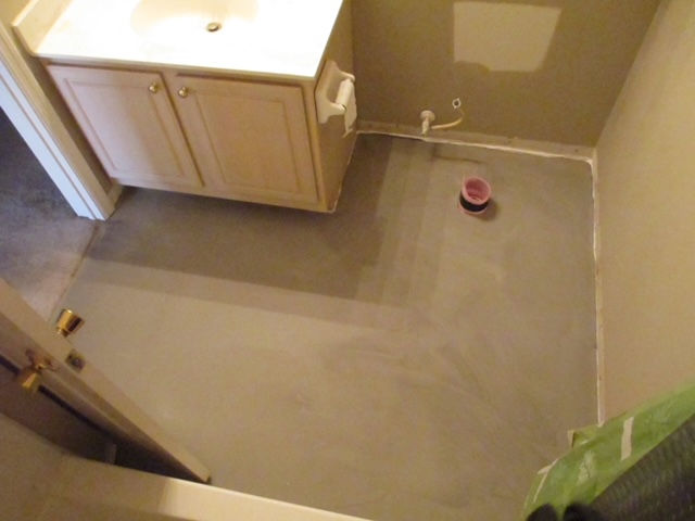 A bathroom under renovation with a newly applied gray floor leveling compound and a prominent toilet drain pipe.