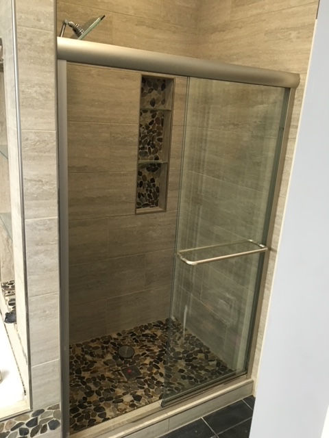 A walk-in shower with a glass sliding door, beige tile walls, a pebble-tiled floor, and a matching stone-tiled shower niche.