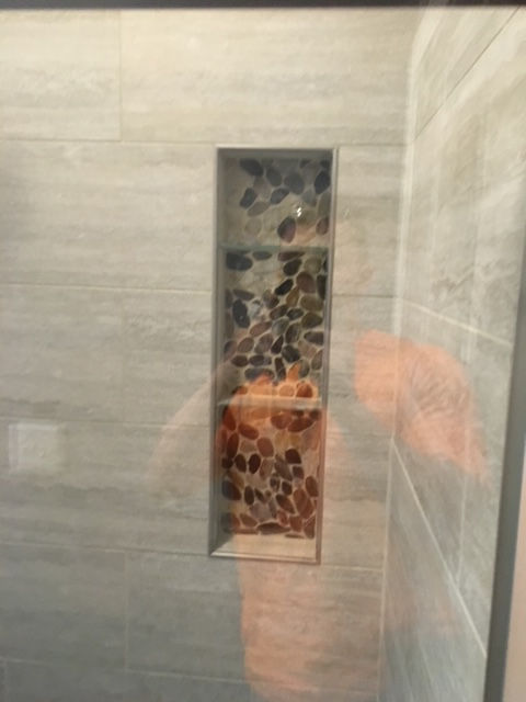 A recessed shower niche featuring multicolored pebble tile lining the back, surrounded by large, light-gray wall tiles.