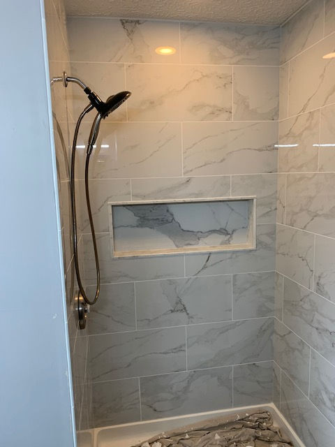 A walk-in shower with white and grey marble-style tiled walls, a built-in recessed shelf, and a black handheld showerhead.