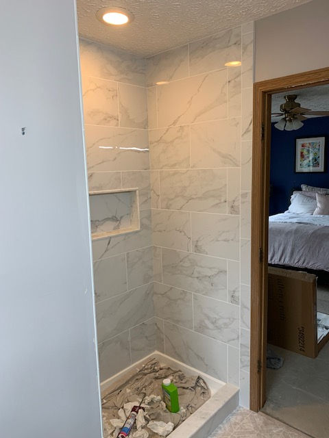A tiled shower stall under construction with a niche, marble-look walls, and installation supplies on the floor.