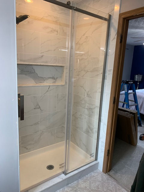 A modern shower stall featuring white marble-patterned tile, a built-in recessed shelf, and a sliding glass door.