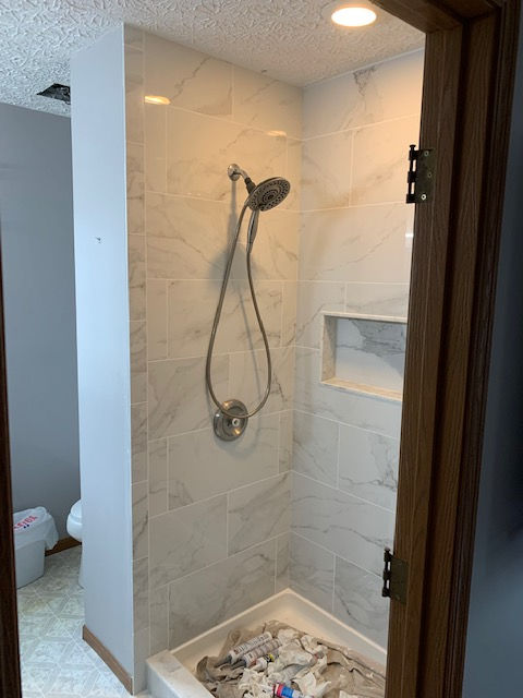 A walk-in shower with white marble-patterned tile, a chrome handheld showerhead, a built-in wall niche, and a white base.