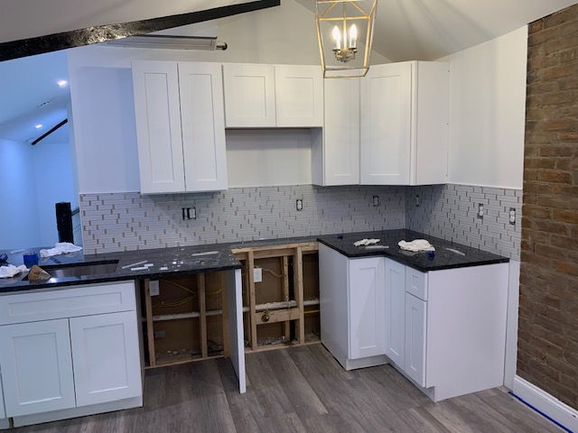 A kitchen under construction with white cabinets, black countertops, and a tiled backsplash, next to a brick wall.