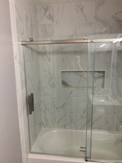 A modern bathtub and shower enclosure with marble-patterned white tiles and a sliding glass door featuring a recessed niche.
