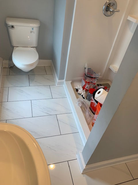A bathroom with a white toilet on marble-style tiled floors and a shower stall containing clutter and toiletries.