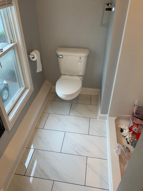 A white toilet centered in a small bathroom with gray walls, light marble-look tile flooring, and a window to the left.