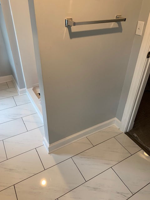 A gray wall with a silver towel bar, adjacent to a shower entrance with white marble-style tiled floors.