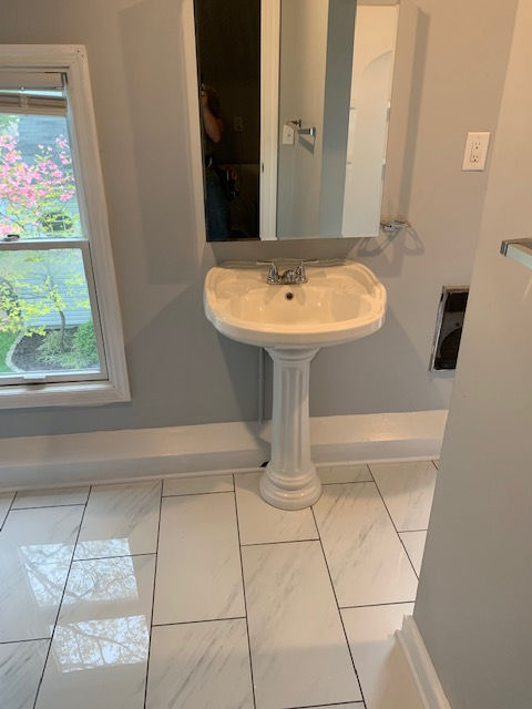 A pedestal sink with a faucet and mirror mounted on a gray wall in a bathroom with white marble-patterned tile flooring.