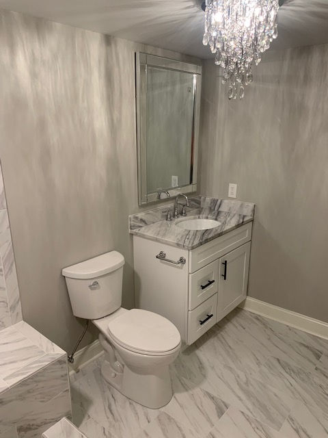 A small bathroom featuring a white vanity with a marble countertop, a mirror, a toilet, and a crystal chandelier.