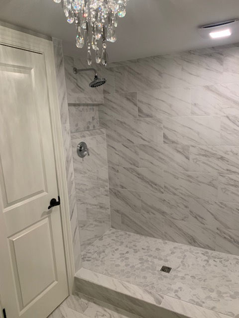 A walk-in shower with white marble-patterned tiles, a chrome shower head, a built-in niche, and a crystal chandelier.