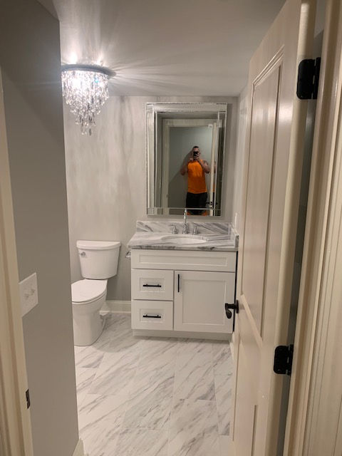 A small, brightly lit bathroom with white cabinets, marble-patterned flooring, and a crystal chandelier.