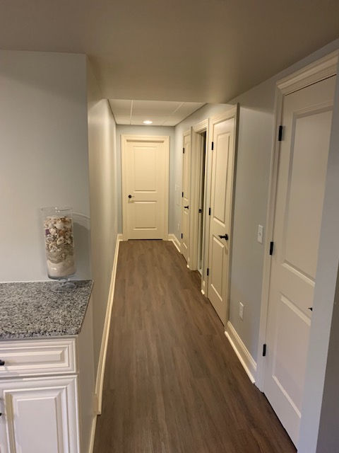 A hallway with light gray walls, brown wood-look flooring, and several closed white doors leading to a room at the end.
