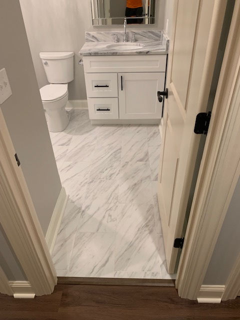 A view through a doorway into a small bathroom with white cabinets, a marble-patterned vanity top, and marble-look flooring.