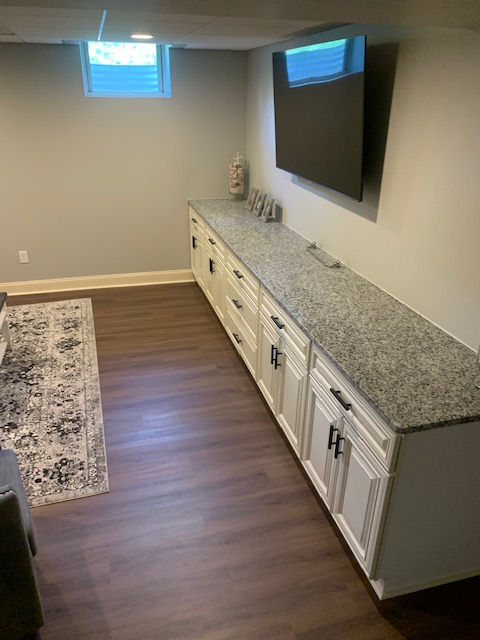 A basement room features a wall-mounted TV above a long, white storage cabinet with a grey granite countertop.