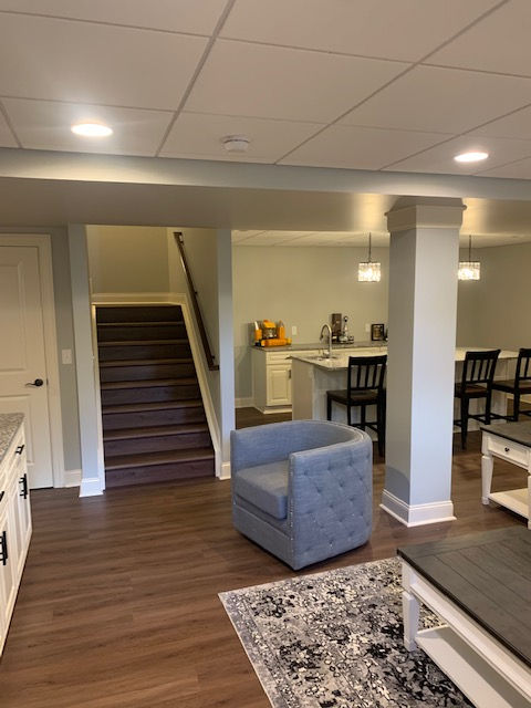 A basement living area with a blue tufted armchair, wood-look flooring, a staircase, and a kitchen island with bar stools.