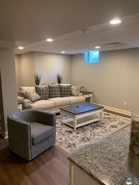 A finished basement with a beige sofa, a gray accent chair, a coffee table on a patterned rug, and recessed lighting.