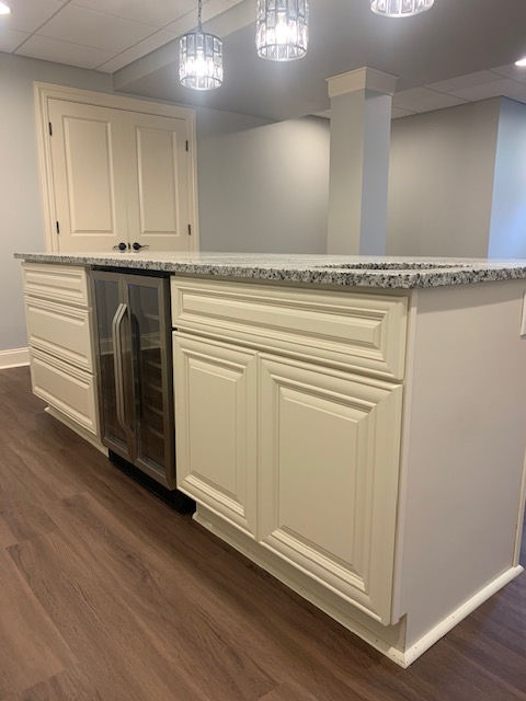 Off-white kitchen island with a wine cooler, gray granite countertop, and light-colored wood flooring in a basement space.