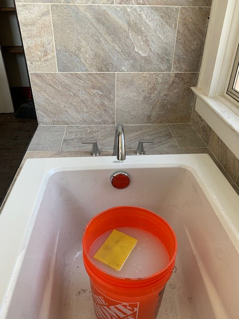 An orange plastic bucket filled with soapy water and a yellow sponge sits inside a white bathtub with tile surrounds.