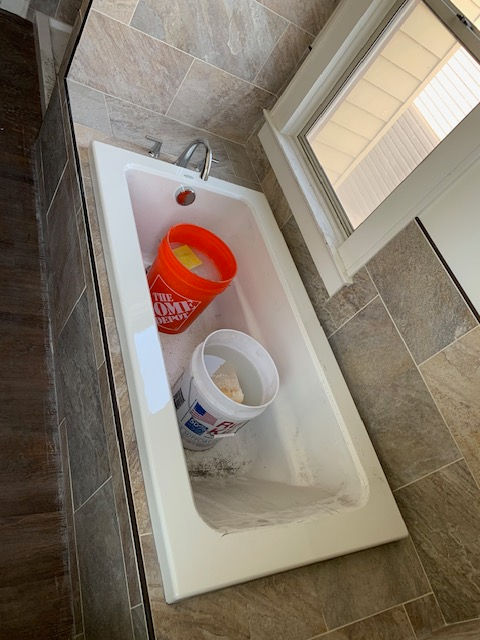 An orange Home Depot bucket and a white bucket sit inside a white bathtub tiled with brown stone in a bathroom.