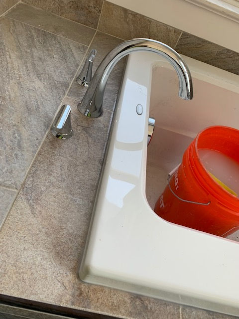 A chrome bathtub faucet with two handles next to an orange bucket sitting in a white tub on a tiled surface.