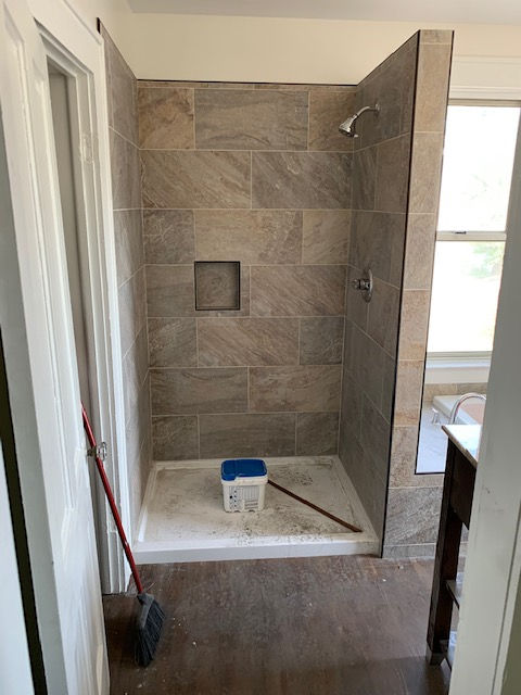 A walk-in shower with brown tiled walls, a built-in niche, a showerhead, a white base, and a bucket inside.