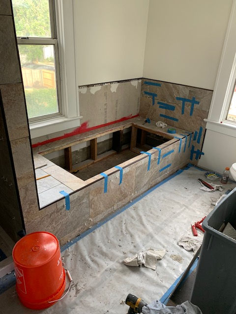 Bathtub area under renovation with wooden framing, partial tiling, and blue painter's tape on beige stone tiles.