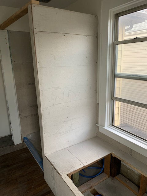 Unfinished plywood shower stall frame and adjacent bench base next to a window in a residential room under renovation.