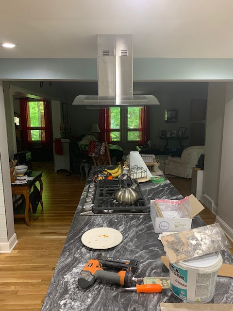 A kitchen island with a dark marble countertop holds construction tools, a burner, and clutter, beneath a metal range hood.
